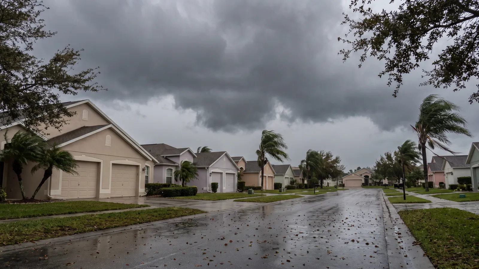 Low storm clouds over a Port St. Lucie street of one-story homes with two-car garages, palms bending and the pavement already wet
